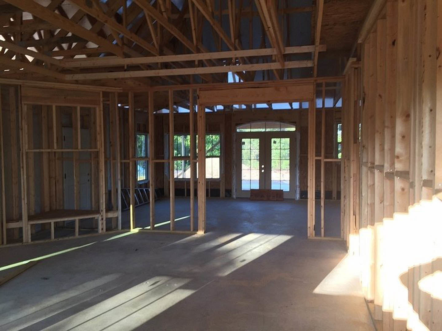 Double glass-paneled doors and a large window illuminate the wooden floor; exposed ceiling beams, a wooden bench with white railing, and sheer white curtains complete the room’s