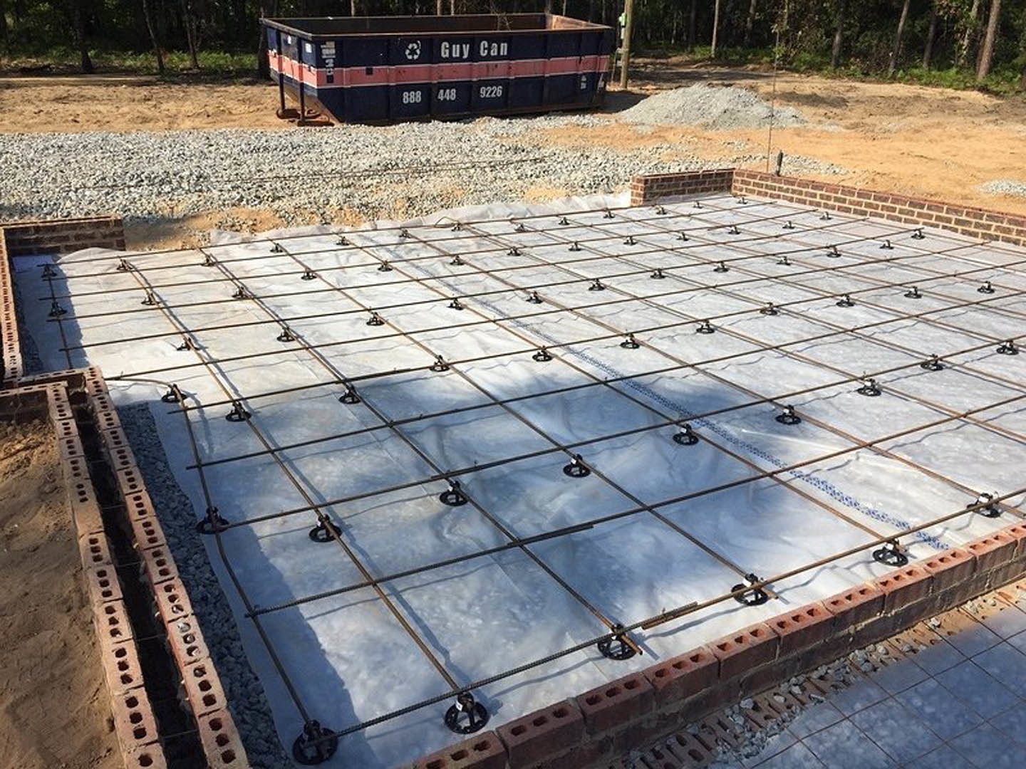 Concrete slab foundation with protruding metal rebar, surrounded by dirt, rocks, and construction materials; garage structure and parked truck visible in background.