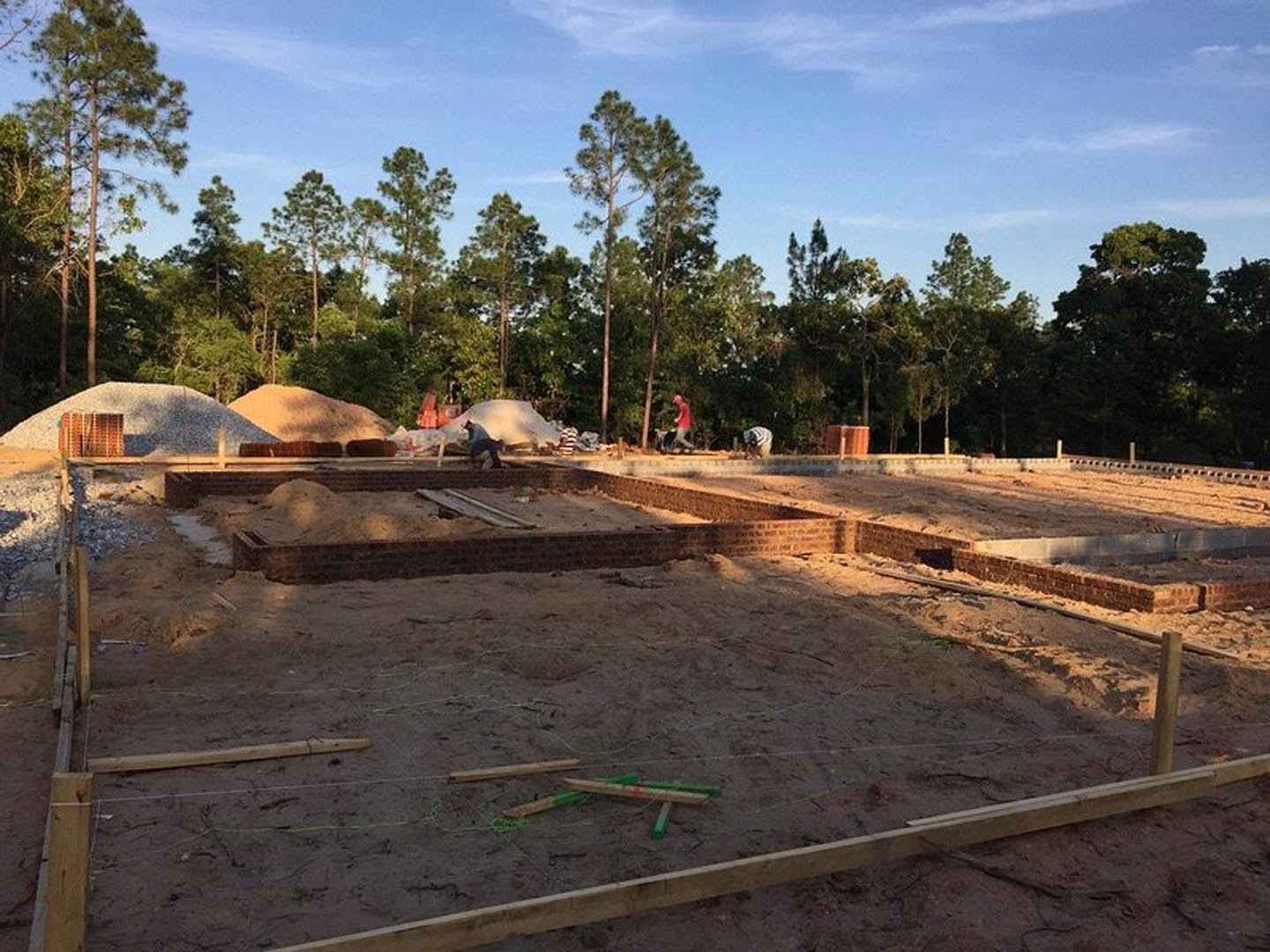 Framed custom home under construction with exposed wooden beams, brick foundation, surrounding trees, and clear blue sky