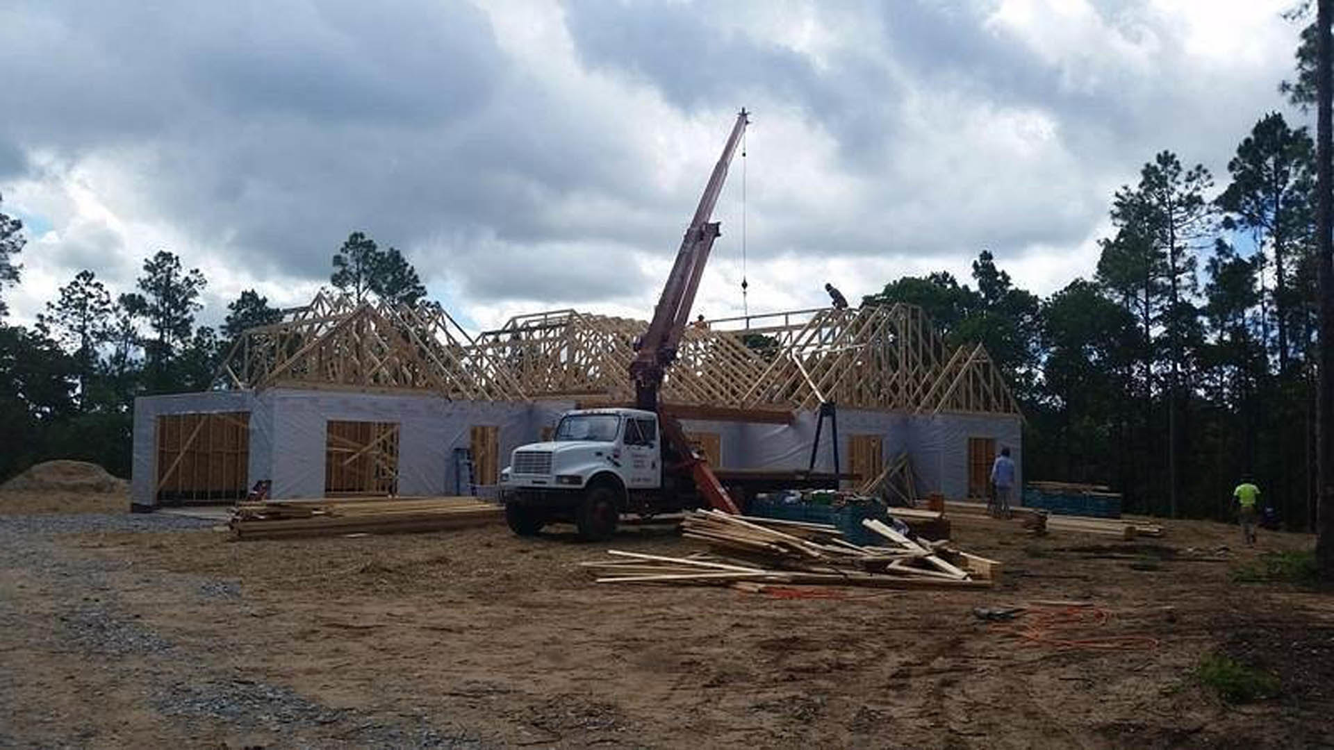 Construction site with a crane lifting materials beside a partially built custom home, white truck parked on dirt, pile of lumber in foreground, barred window visible on house
