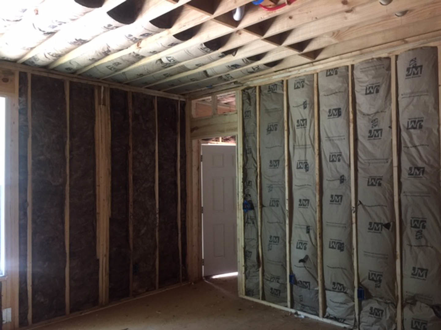Room with unfinished wood panel walls, white door featuring square pattern, exposed insulation covered by white sheets, shelf holding newspapers, plywood ceiling beams.
