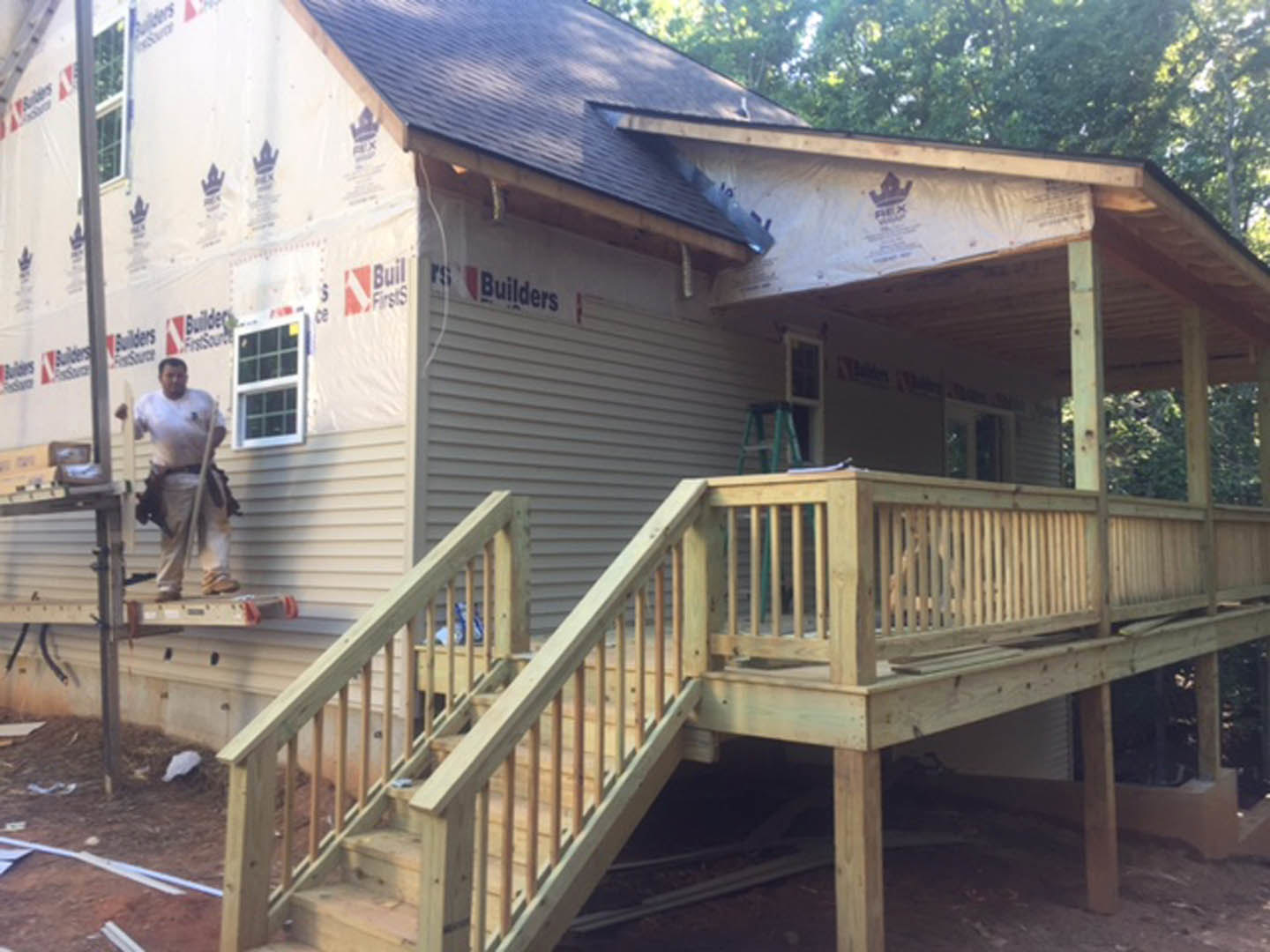 Wooden porch with railings, man in casual clothing standing near white siding, multiple square windows, gabled roof, trees and fence in background