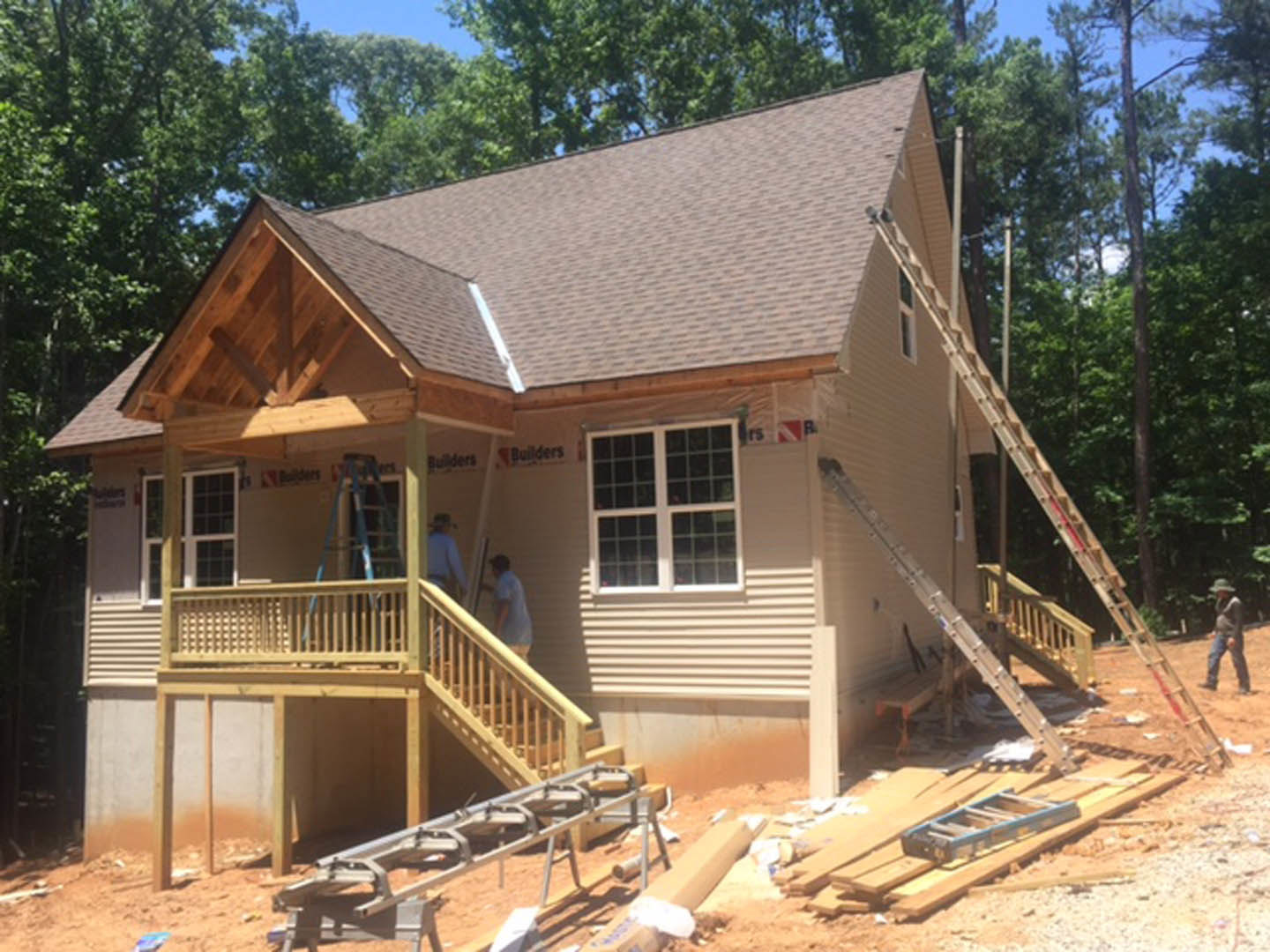Wood-framed house under construction with exposed lumber, ladders leaning against exterior walls, unfinished porch, and large windows; trees and sky visible in background.