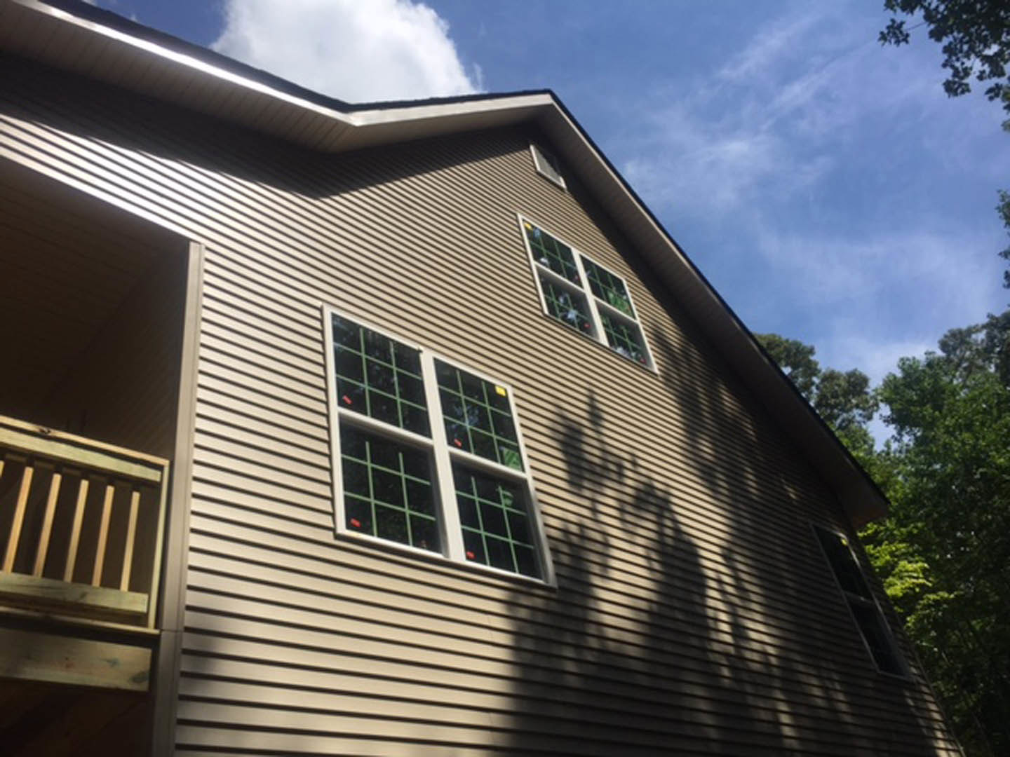 Modern home exterior featuring numerous large windows, light-colored siding, wooden porch railing, and trees in the background under a clear blue sky