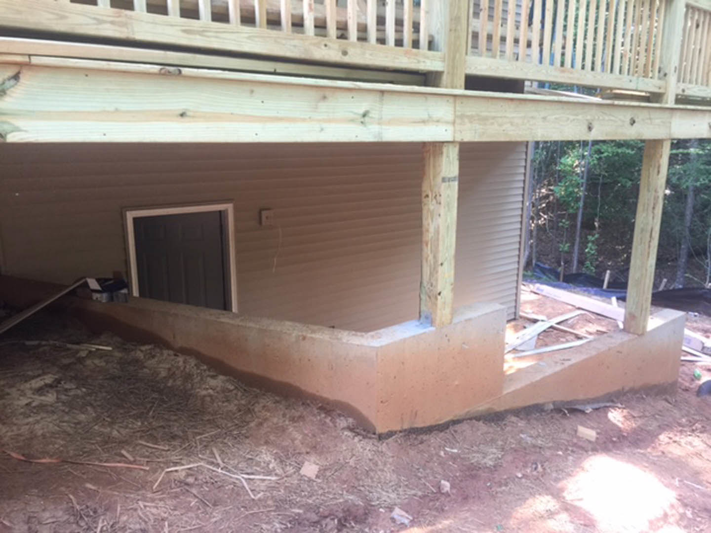 Partially built house with exposed wooden deck and stairs, concrete foundation, scattered construction materials on dirt ground, unfinished exterior walls, and visible window