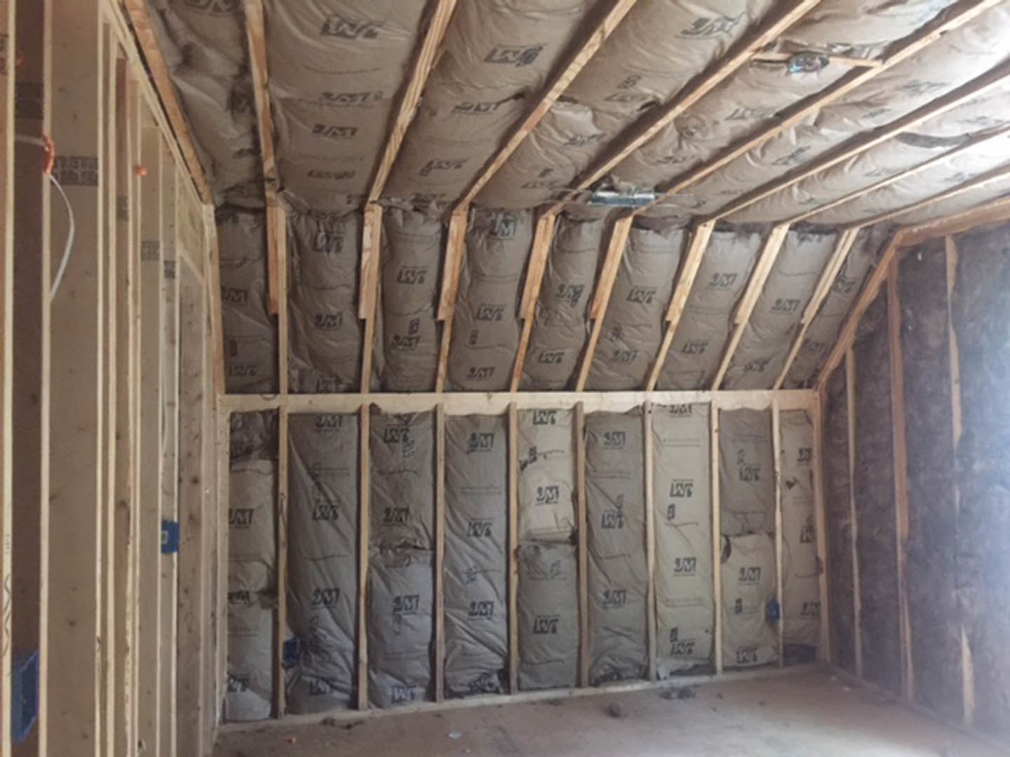 Room under construction featuring exposed wood beams, a wall lined with black insulation bags, and a ceiling with white insulation material; grey construction equipment rests on