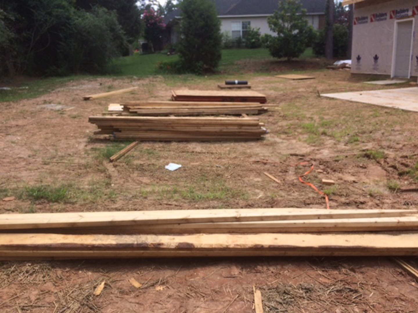 Stack of lumber planks on grassy yard beside white house with door, tree trunk visible in background