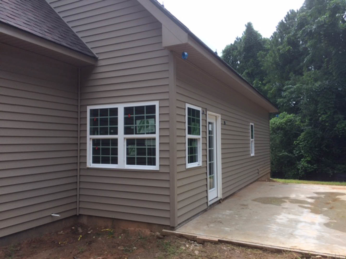 Modern home with light-colored siding, white-framed windows, wet concrete patio, and mature trees in the background