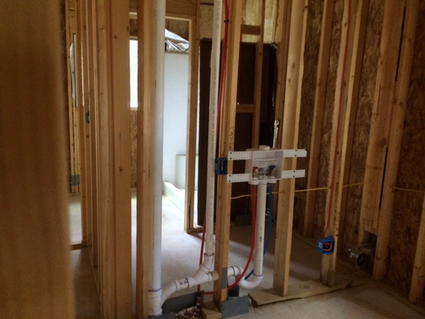 Unfinished room with exposed wood framing, white PVC pipe, red and blue PEX tubing, electrical wires, plywood subfloor, and construction materials