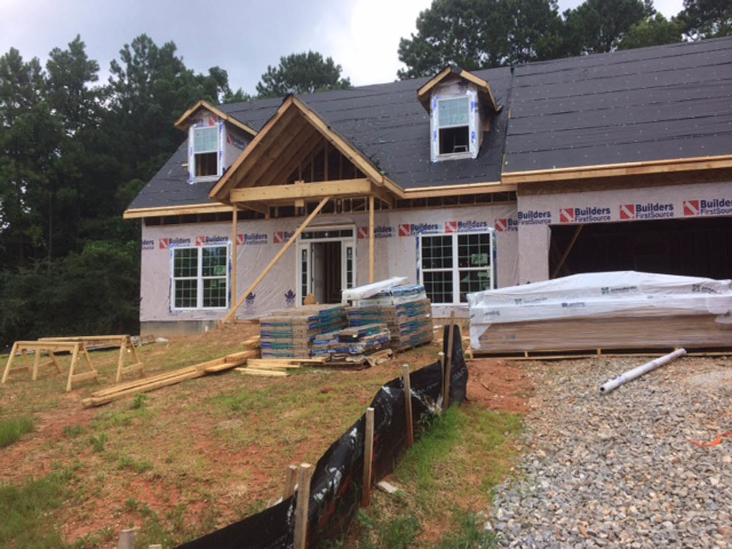 Framed house under construction with exposed wooden beams, roof partially completed, piles of lumber and cardboard boxes on the ground, white plastic bag and pipe near foundation