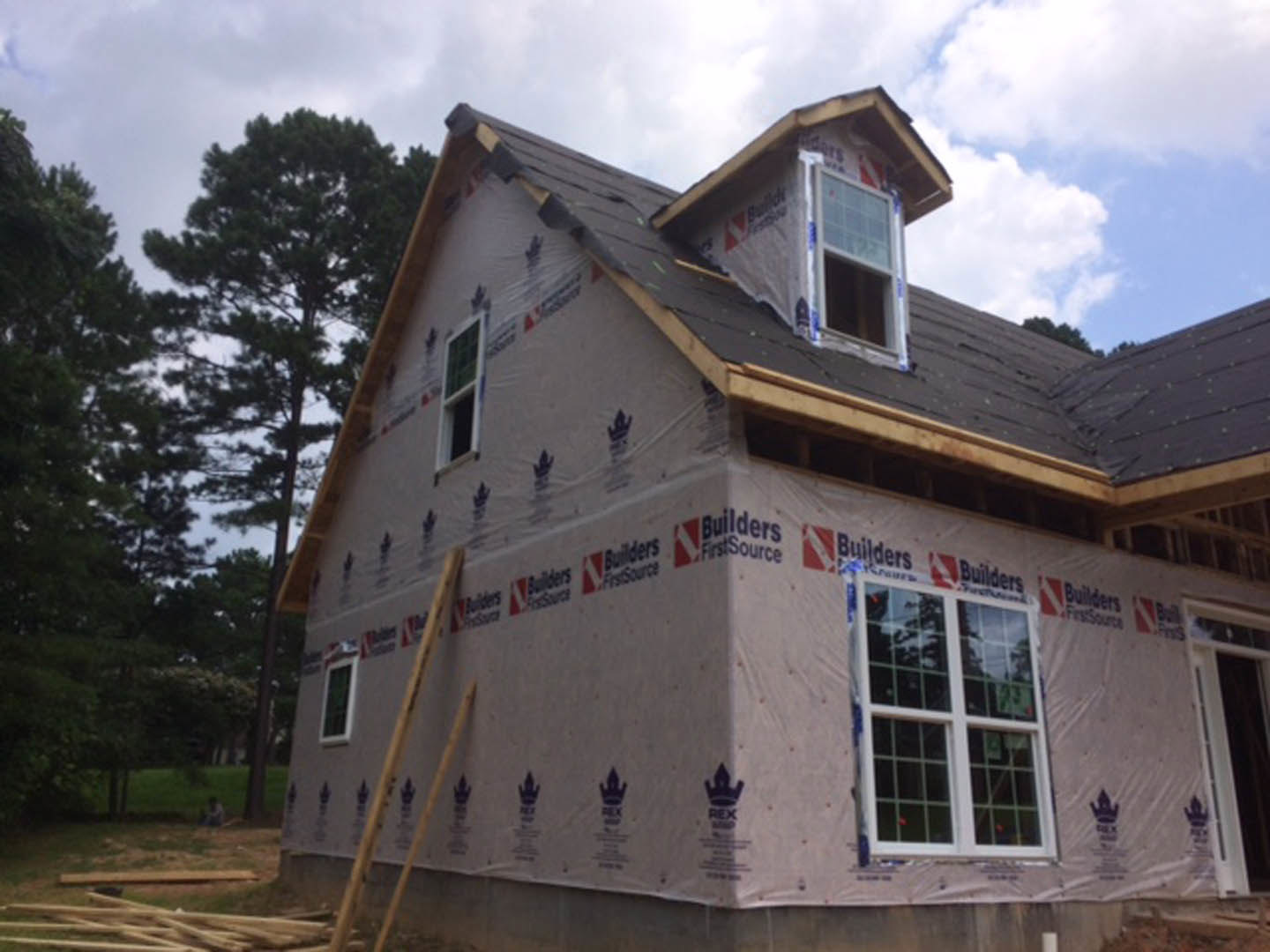Partially built house with exposed framing, unfinished roof, and surrounding trees beneath a clear blue sky