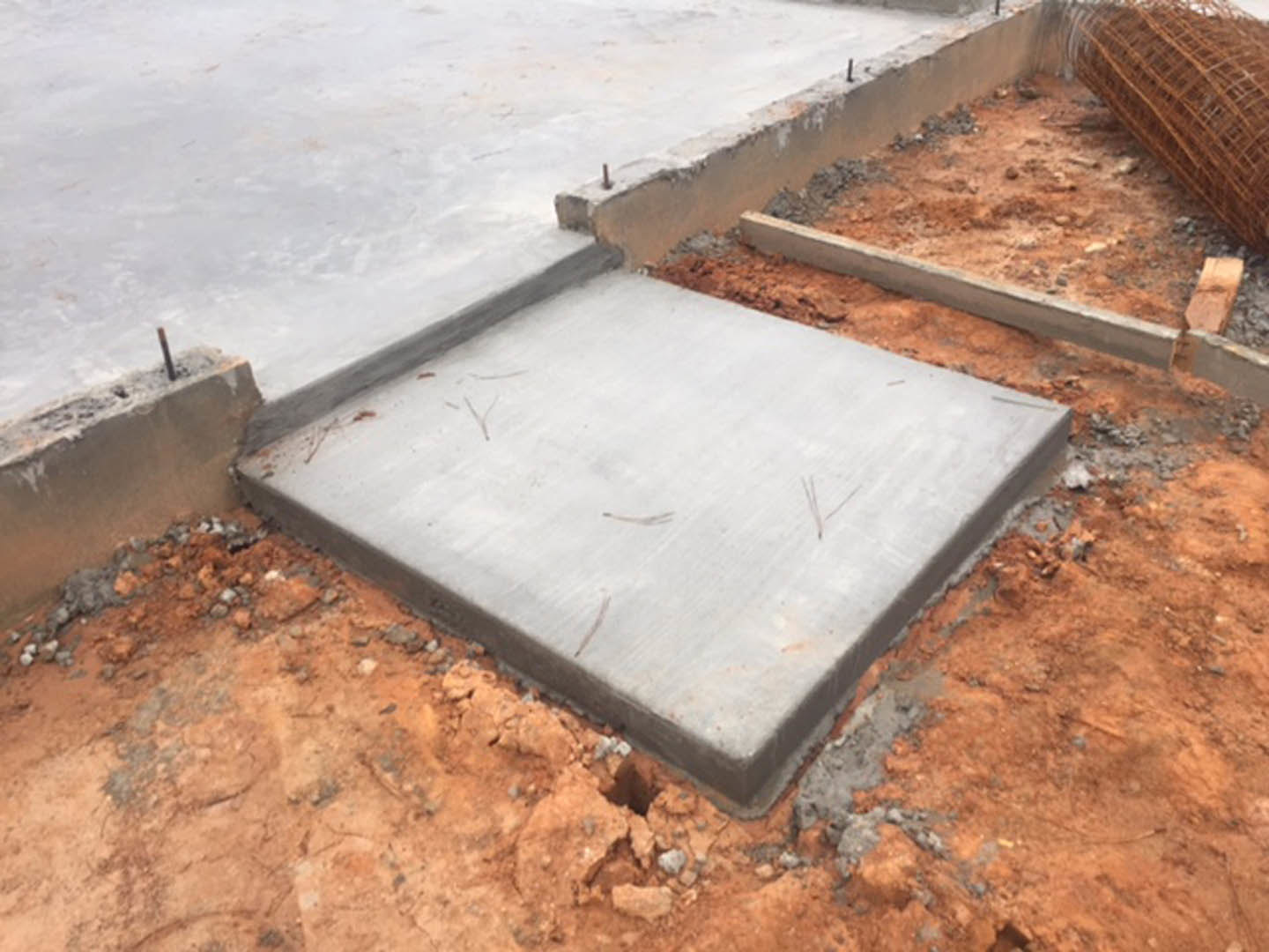 Concrete foundation slab with embedded metal rods and wire mesh, surrounded by soil and construction materials.