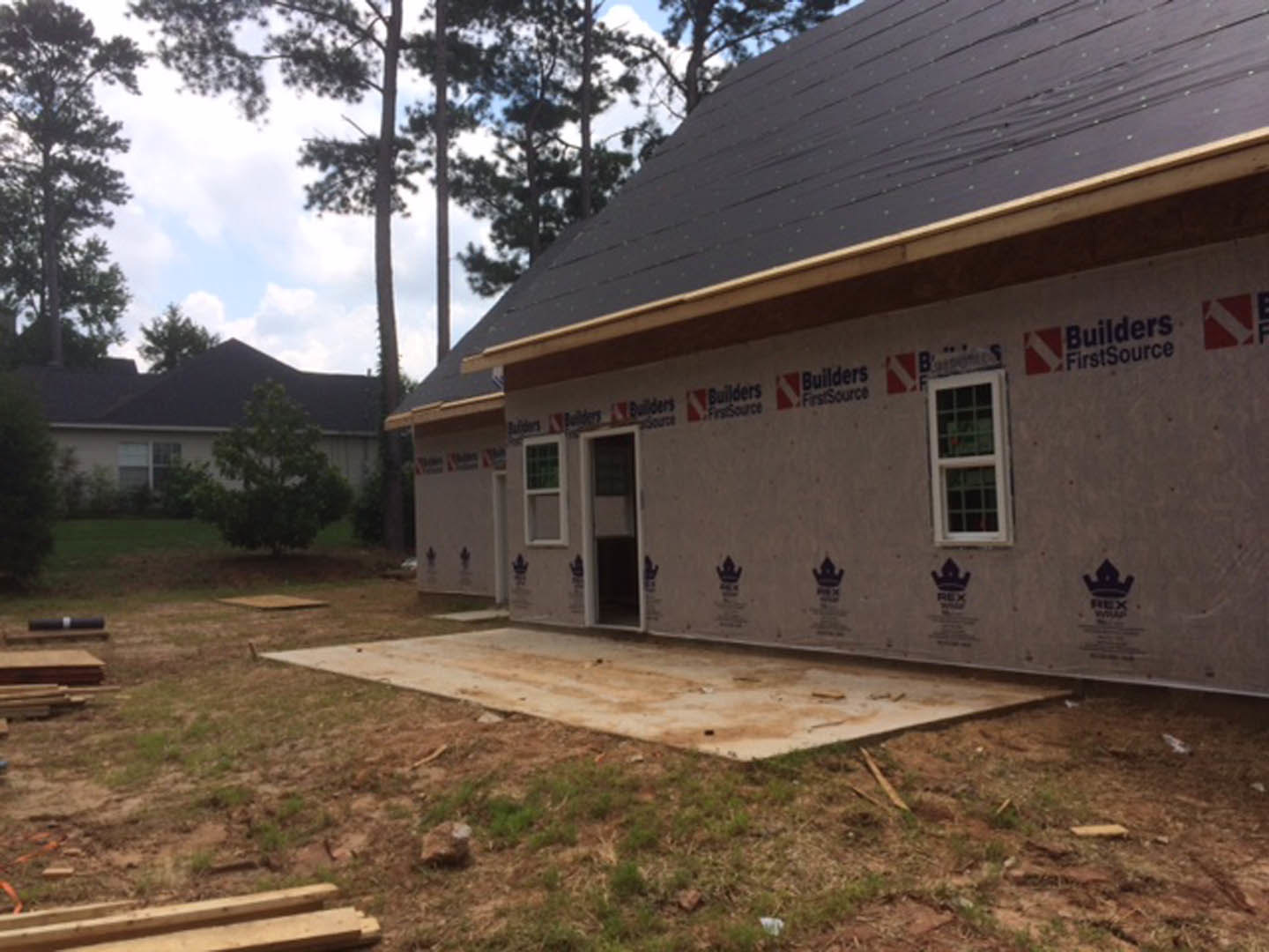Framed house under construction with exposed lumber, multi-pane windows, grassy yard, and surrounding trees