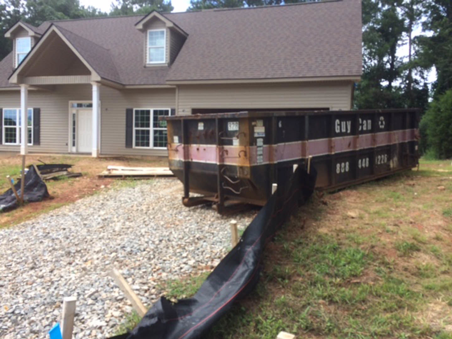Black metal dumpster with white numbers sits on driveway in front of suburban house with brick exterior and large windows, surrounded by grass and trees.
