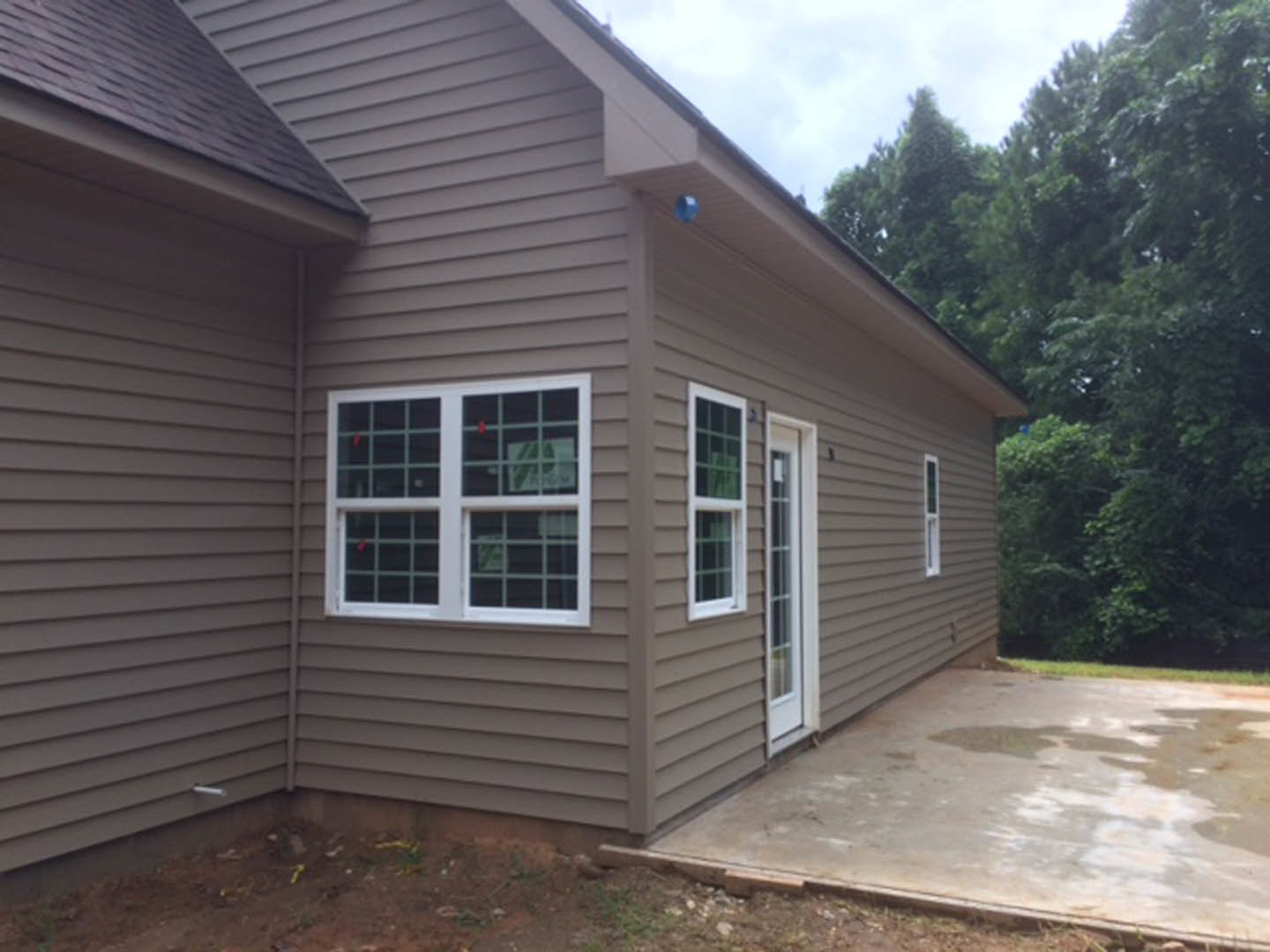Modern house with light-colored siding, white-framed window, wet concrete patio, and mature trees in the background
