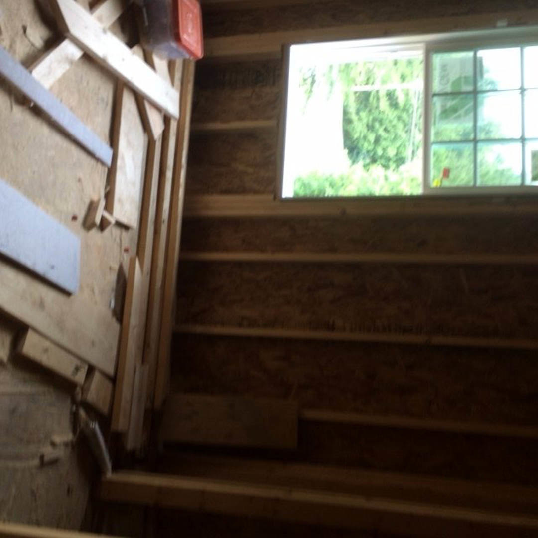 Bright room featuring two white-framed windows, natural daylight, pale painted walls, and stacked lumber visible in the foreground.