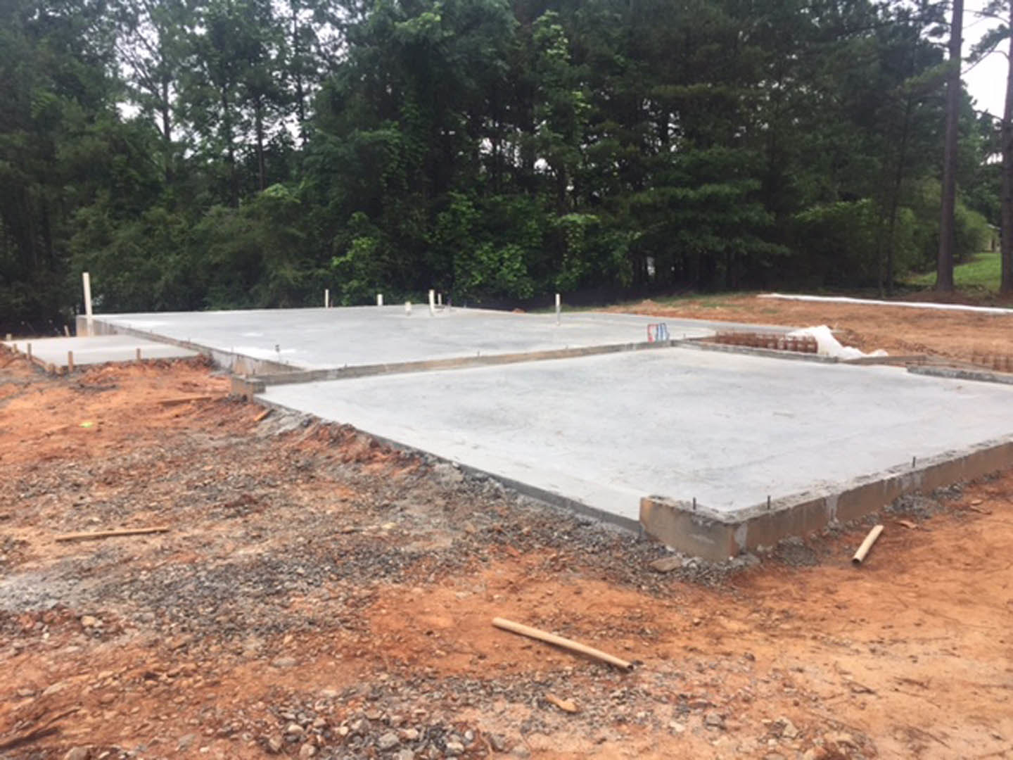 Freshly poured concrete slab foundation on a residential construction site, surrounded by dirt, wooden stakes, and nearby trees under a clear sky.