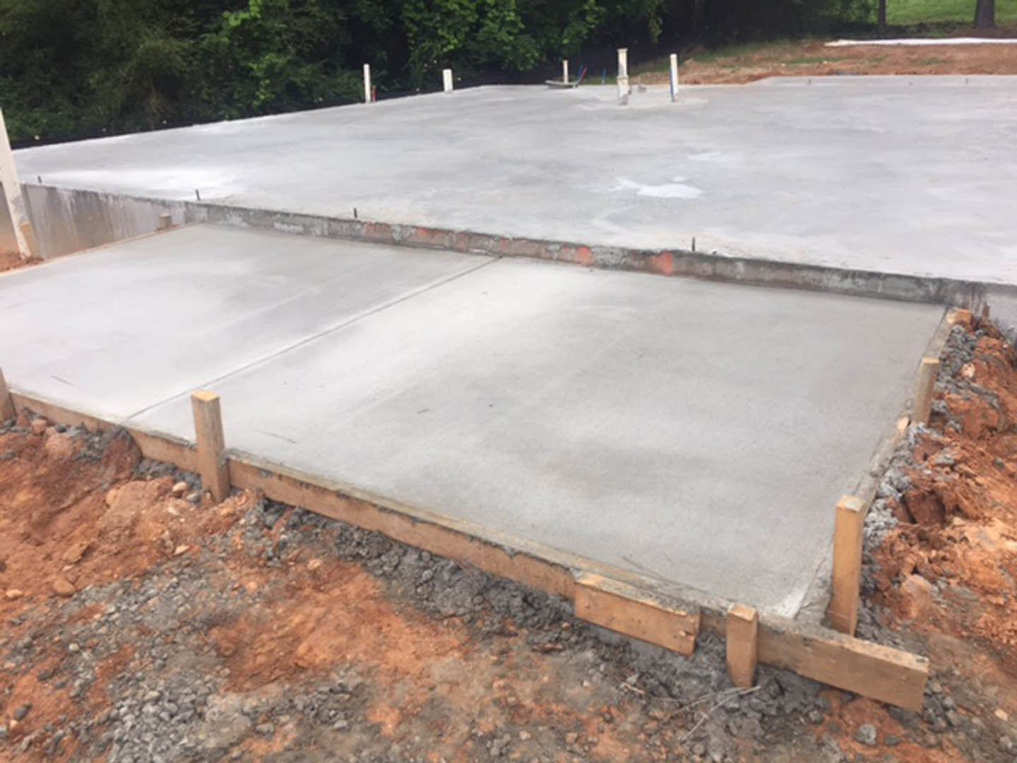 Freshly poured concrete slab foundation with wooden posts, surrounded by outdoor trees and construction materials