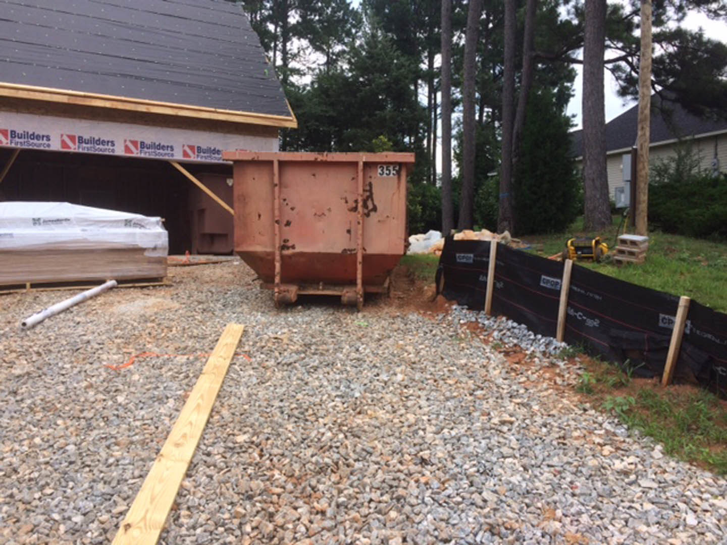 Large metal dumpster on gravel driveway in front of a building, black fabric construction fence with white text, wood planks and lumber pile covered in plastic, trees in