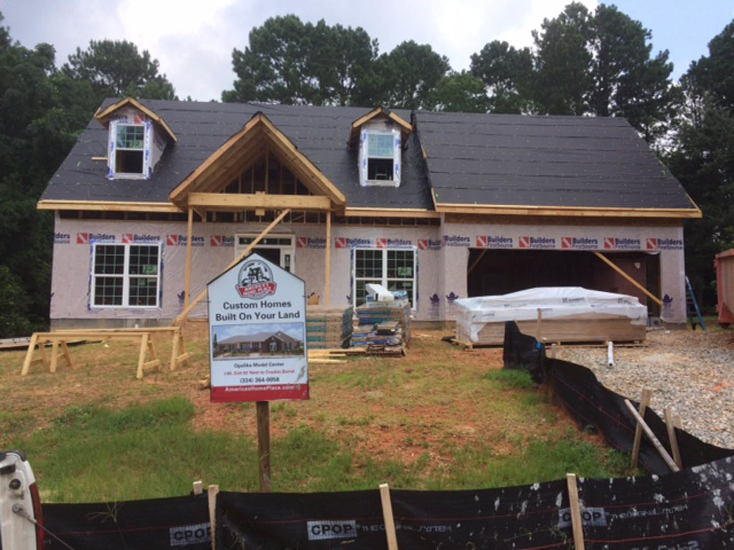 Partially built house with exposed wooden framing, construction sign featuring a house illustration, white siding, black-framed window, and tree in background under blue sky
