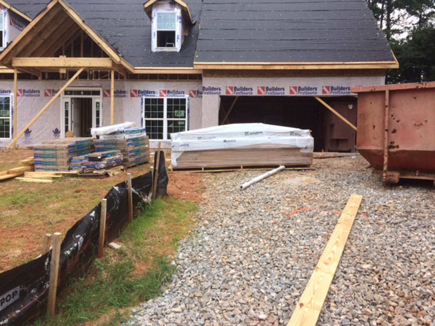 Partially built house with exposed framing, pile of wooden pallets, white plastic bag, black plastic-covered fence, perforated metal container, wood plank on gravel, white-framed