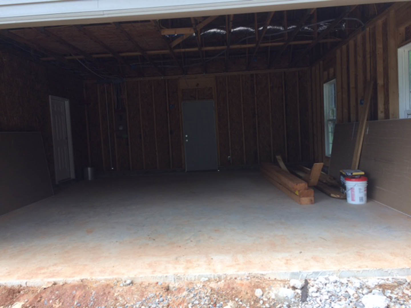Wood planks stacked on a light-colored floor beside a white bucket with red lid, dark wooden door with small dot details, neutral walls in a residential interior.