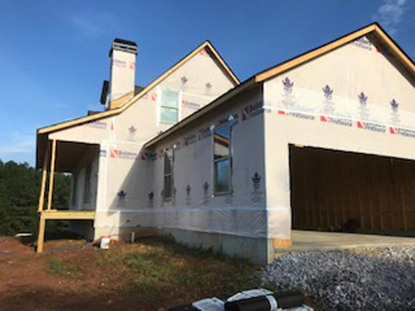 Two-story house under construction with exposed framing, attached garage, white siding partially installed, blue sky overhead
