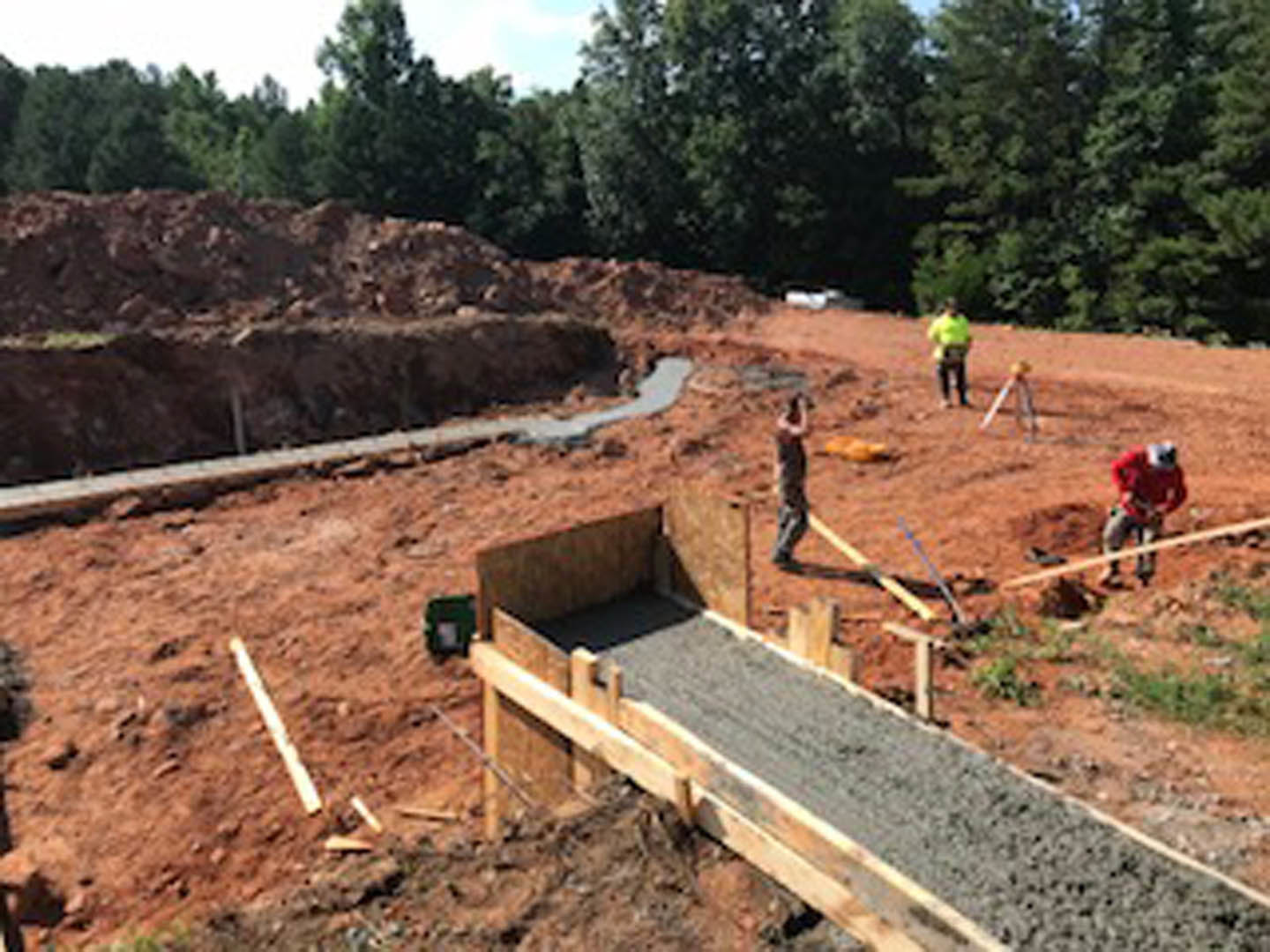 Framed wooden beams and cement foundation at a residential construction site, workers in safety gear, soil and trees in background under clear sky