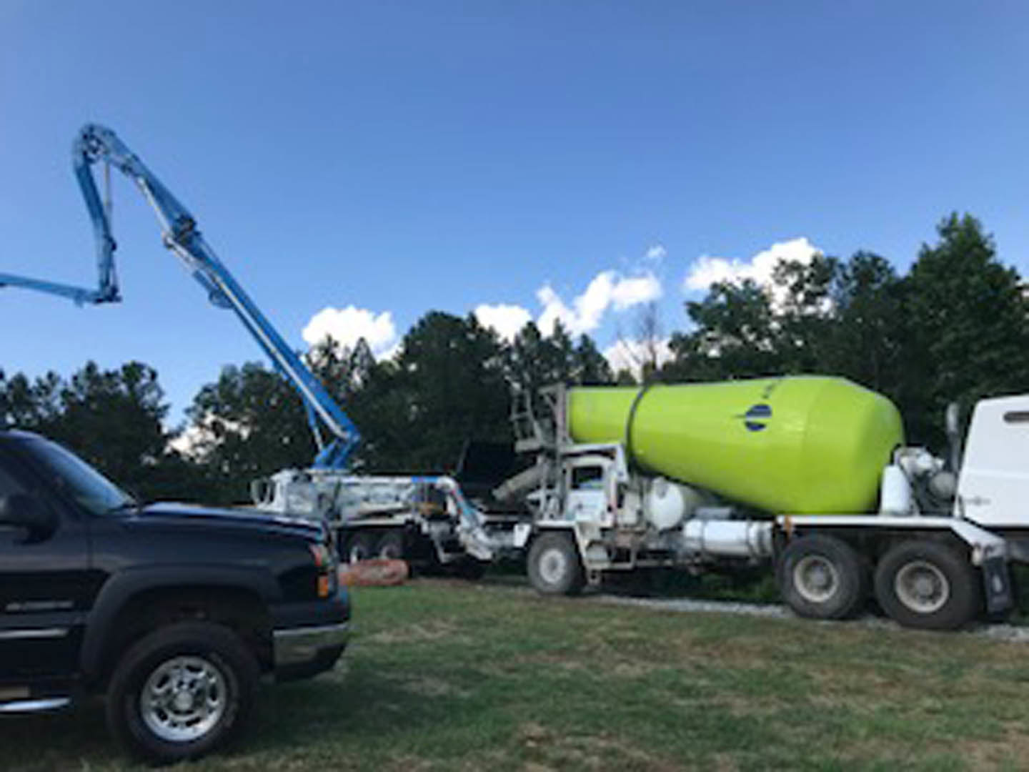 Black truck parked on grass with large green cement mixer attached, blue logo visible on mixer, trees and sky in background