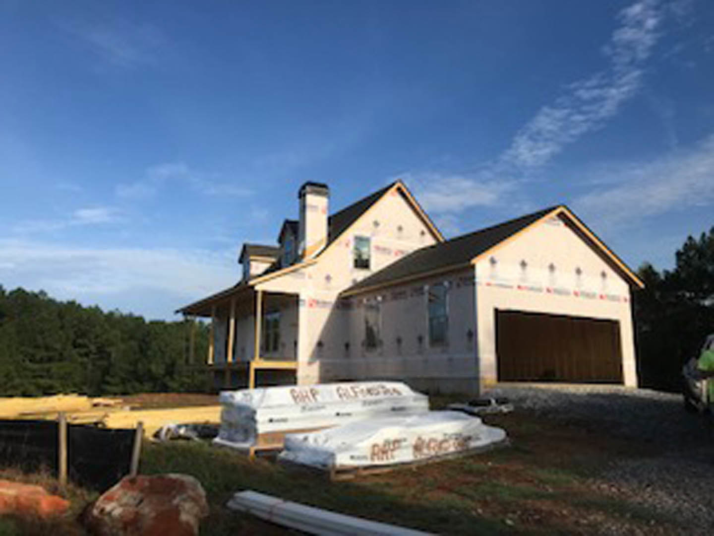 Framed house under construction with exposed wooden beams, stacks of building materials on dirt lot, partially finished chimney, cloudy sky, and surrounding trees