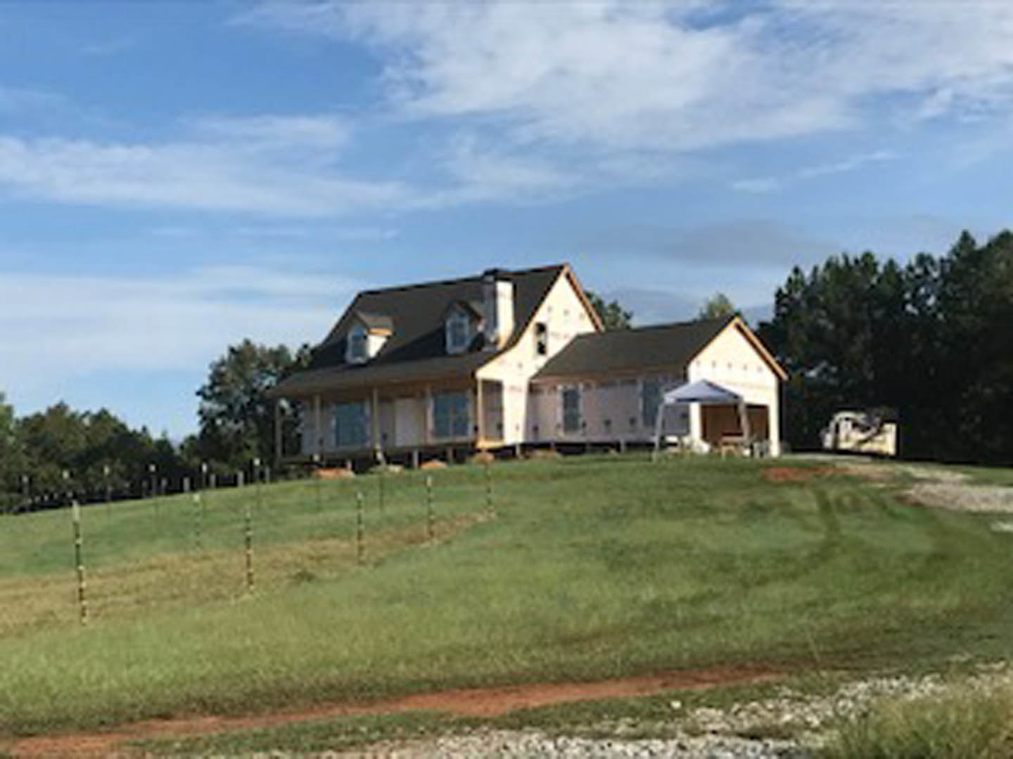White farmhouse with brick chimney sits atop a grassy hill, surrounded by a wooden fence, under a blue sky with scattered clouds.