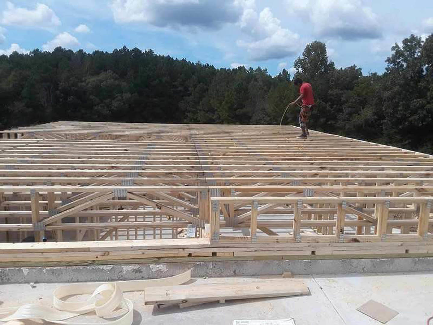 Man in red shirt standing atop partially constructed wooden and metal-framed home, blue sky and clouds above forested landscape, wood flooring and concrete surface visible.