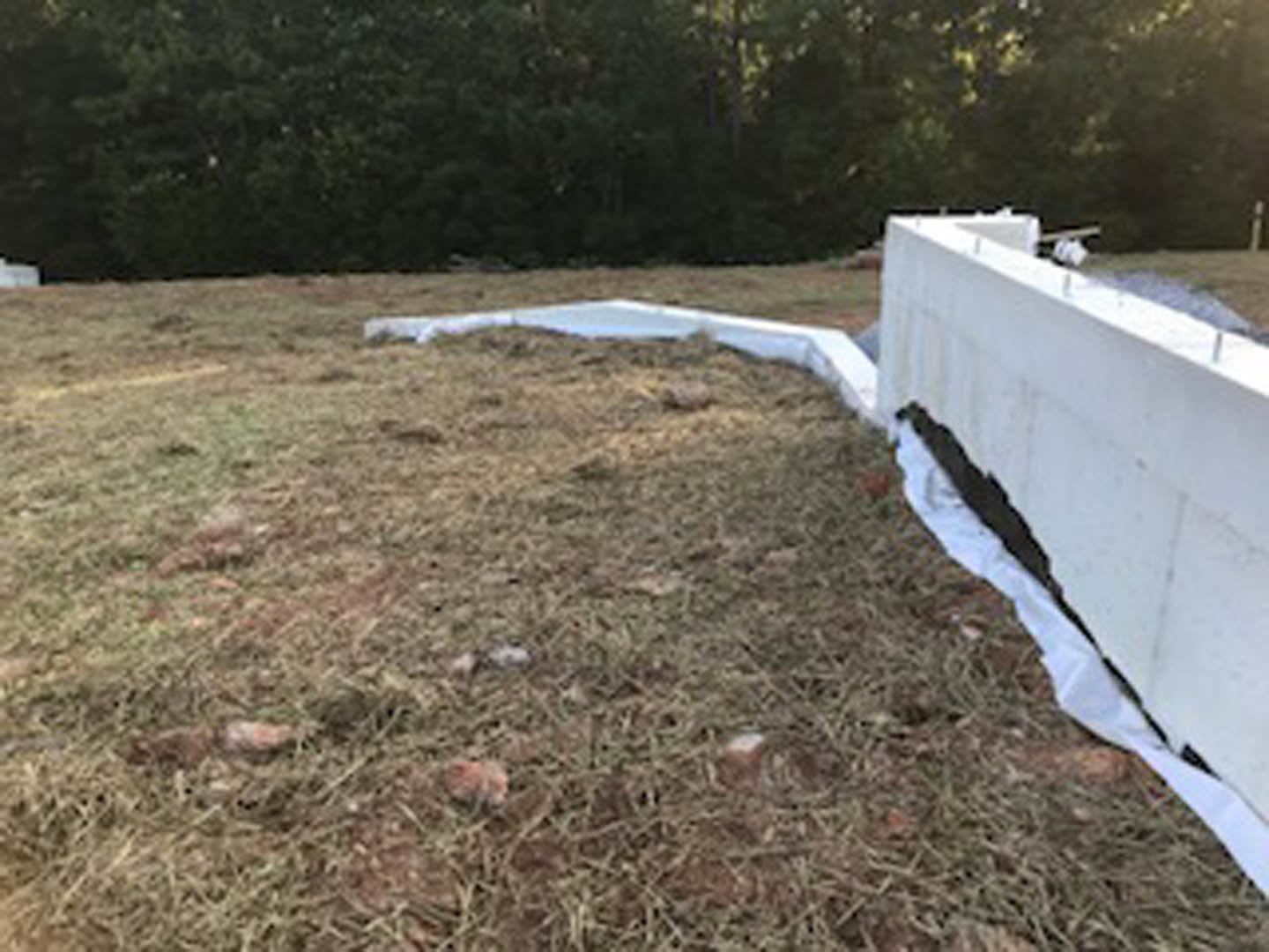 White wall with a white plastic sheet covering, grassy field with scattered rocks in foreground, group of trees in background.
