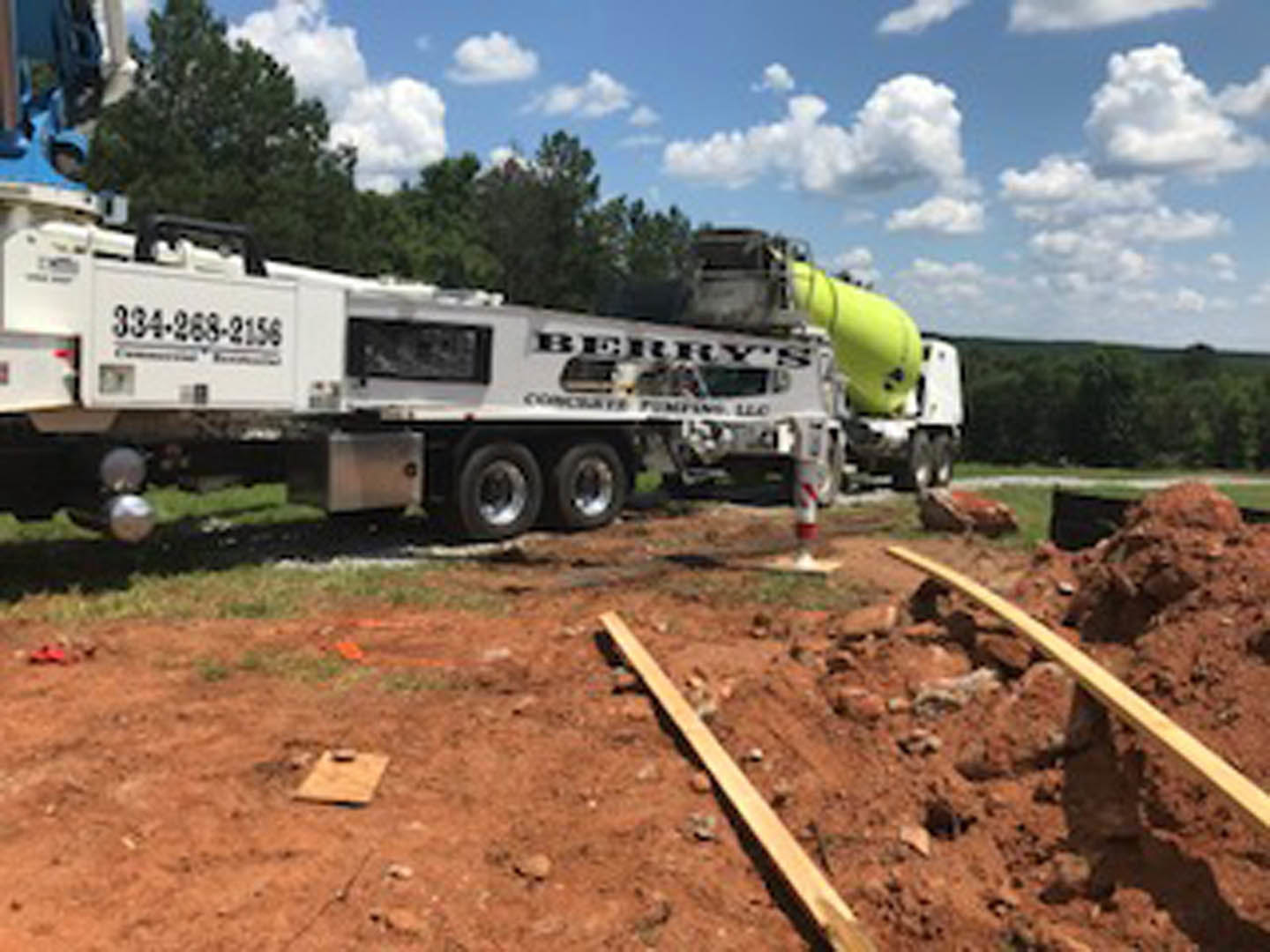 White cement mixer truck with black lettering parked beside a pile of dirt and wood planks, yellow tube mounted on top, blue sky with scattered clouds overhead, partial view of a