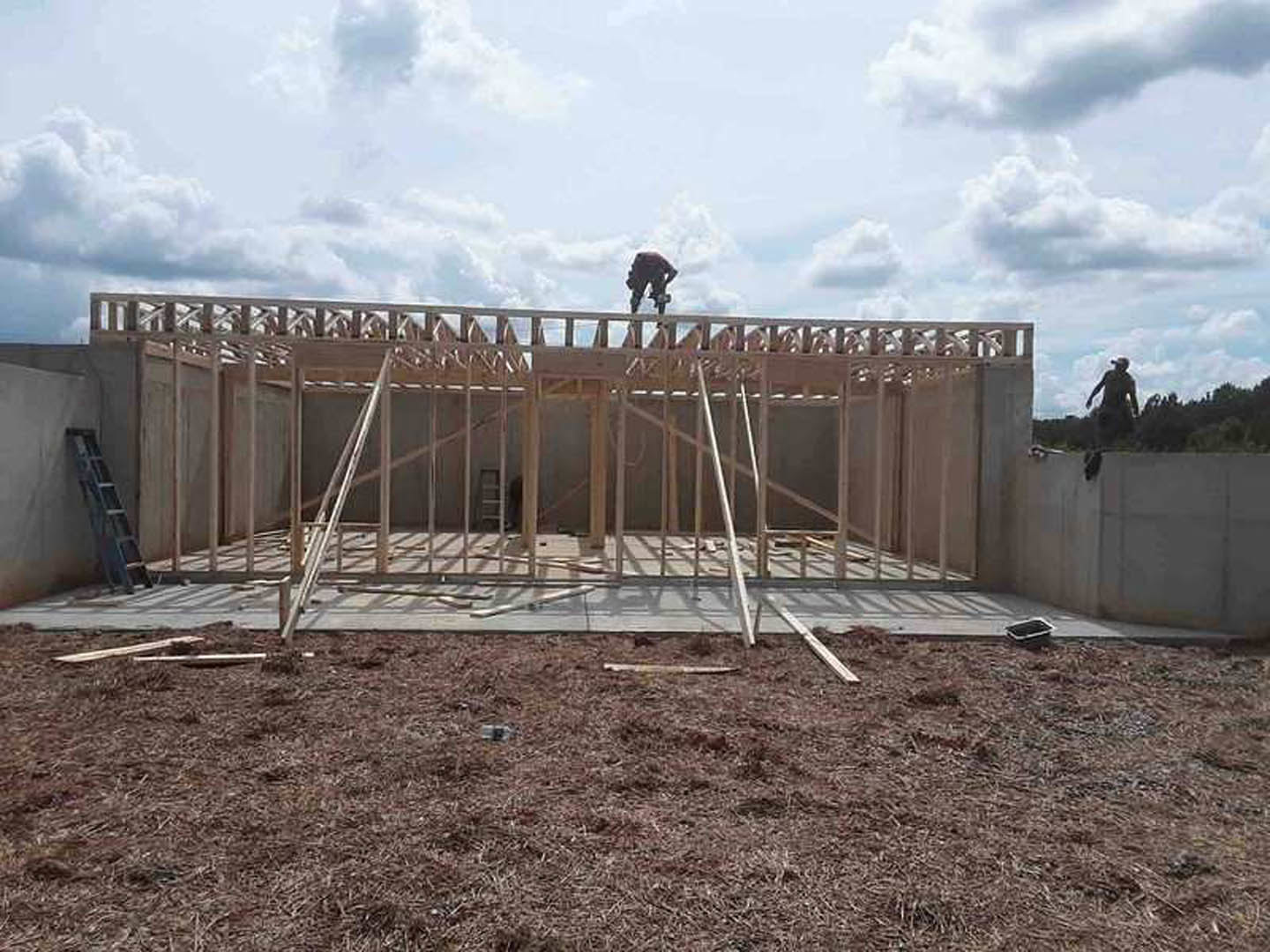Wood-framed custom home under construction with exposed composite materials, foundation, and walls; man standing on roof against cloudy sky.