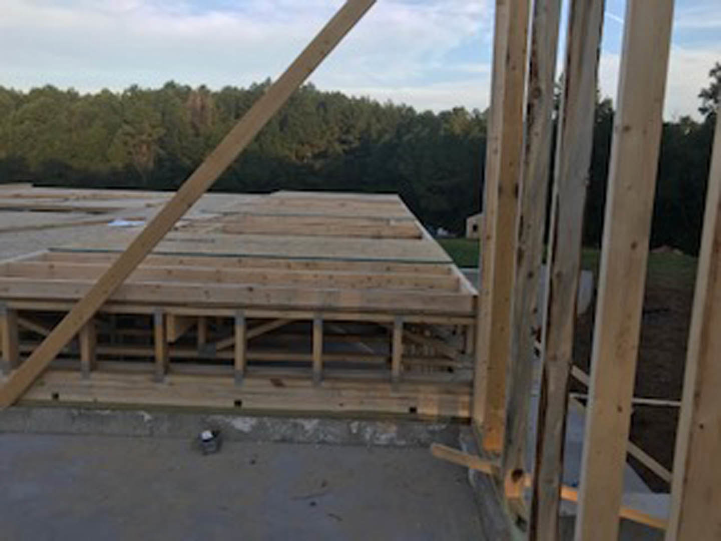 Wood-framed exterior structure under construction with exposed lumber beams and planks, set against a partly cloudy sky and trees in the background