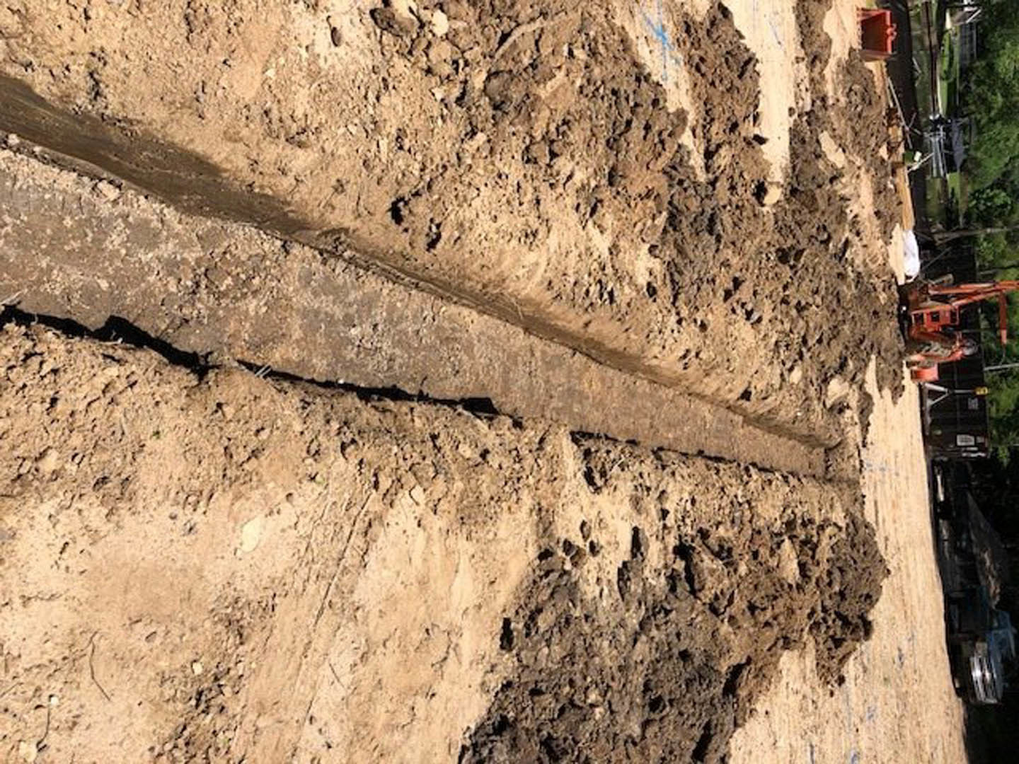 Freshly dug trench in brown soil bordered by loose dirt and small plants, outdoor construction site with trees in background