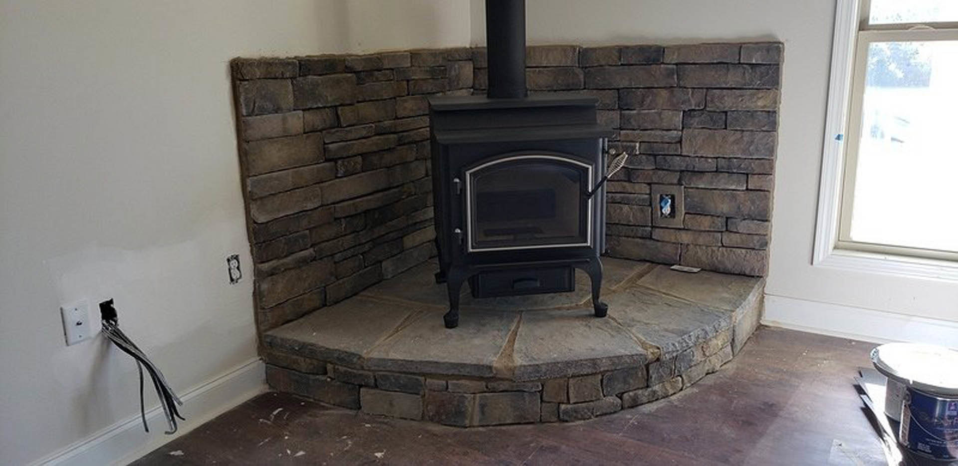 Black wood-burning stove with glass door set against a brick and tile hearth, stone wall backdrop, hardwood floor, and nearby chair in a cozy indoor living space