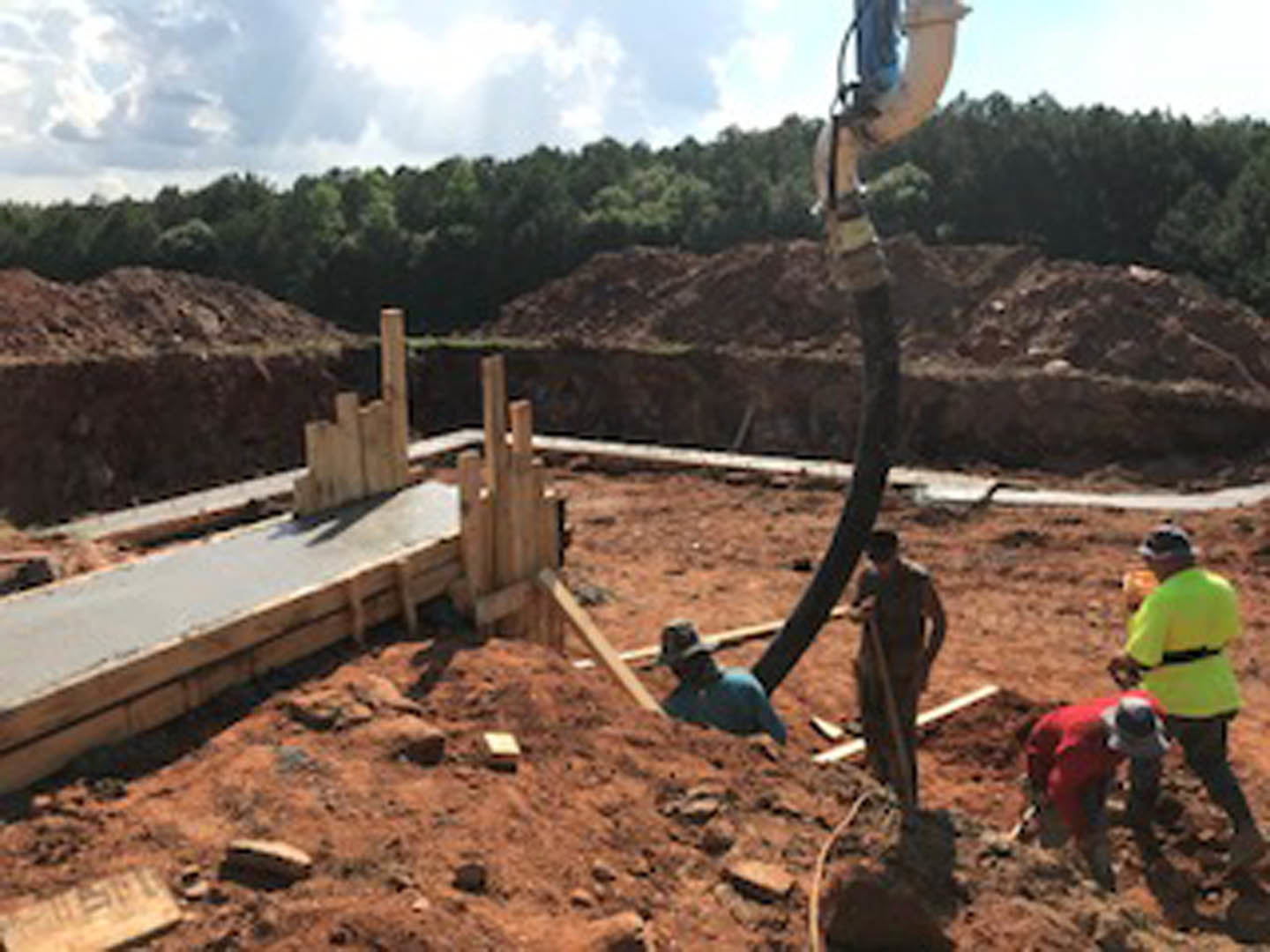 Workers in safety vests and hard hats building a house foundation on a dirt lot, surrounded by construction materials and equipment under a partly cloudy sky