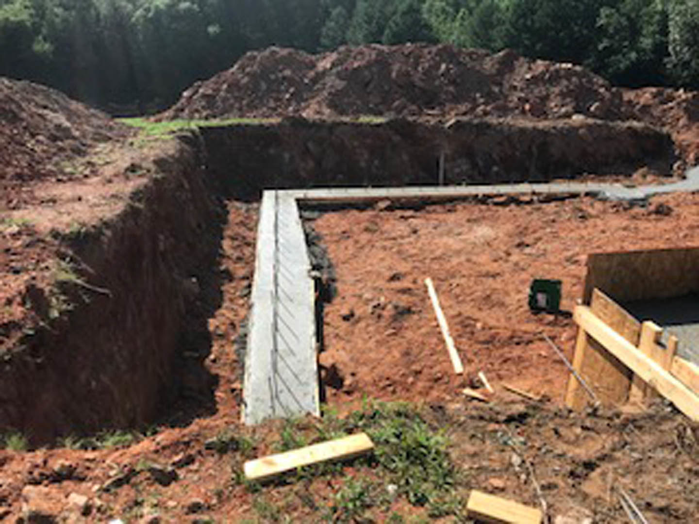Wooden bridge spanning a dirt foundation at a residential construction site, surrounded by soil, scattered wood planks, and nearby trees.