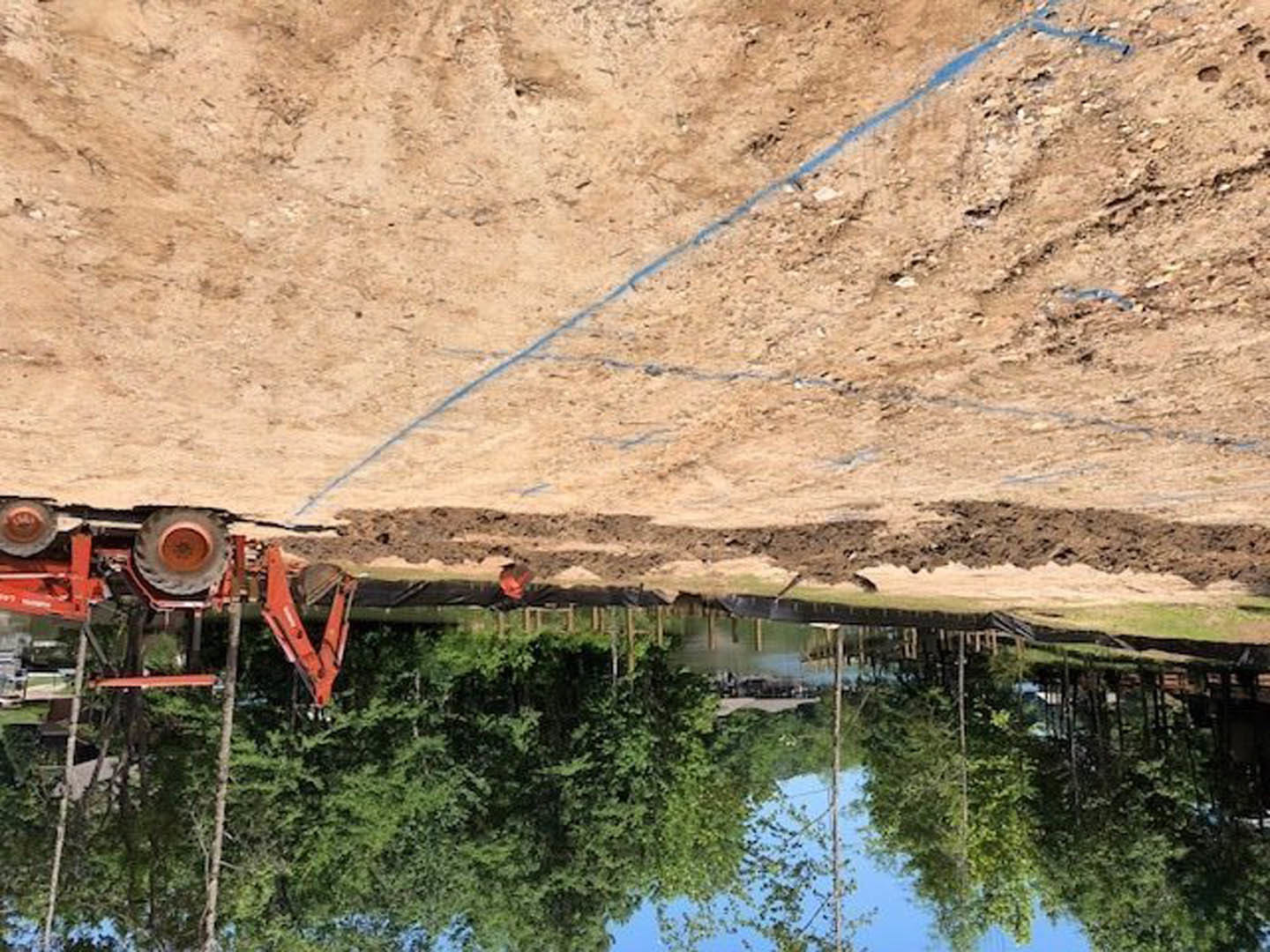 Tractor parked on a dirt road beside a lake, surrounded by trees and water reflections