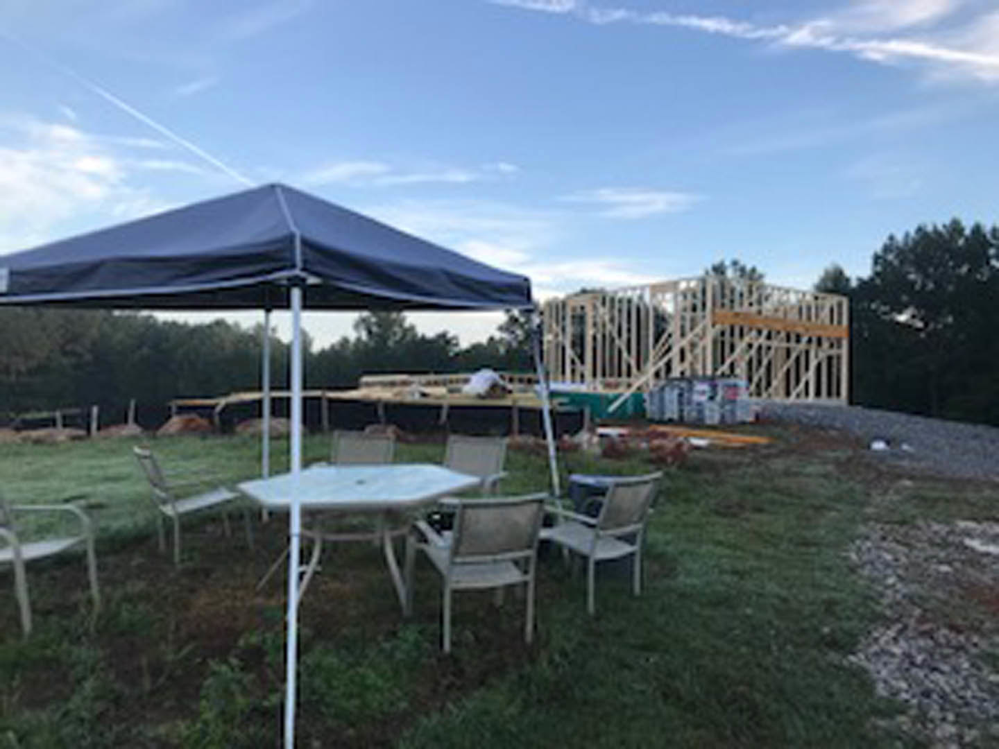 Wooden table and chairs set on green grass beneath a canopy, surrounded by trees and clouds in the sky, outdoor construction materials visible