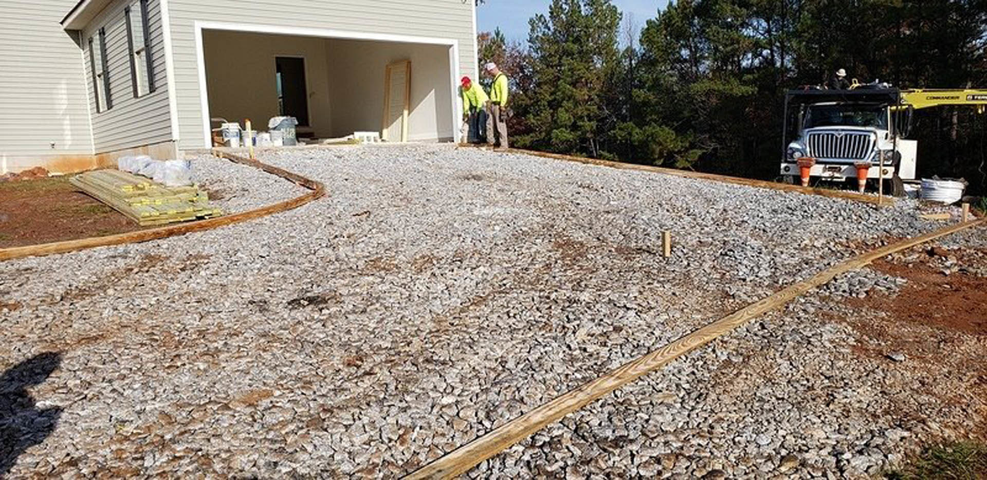 Men standing on asphalt driveway in front of custom home exterior with trees and plants, white truck parked nearby