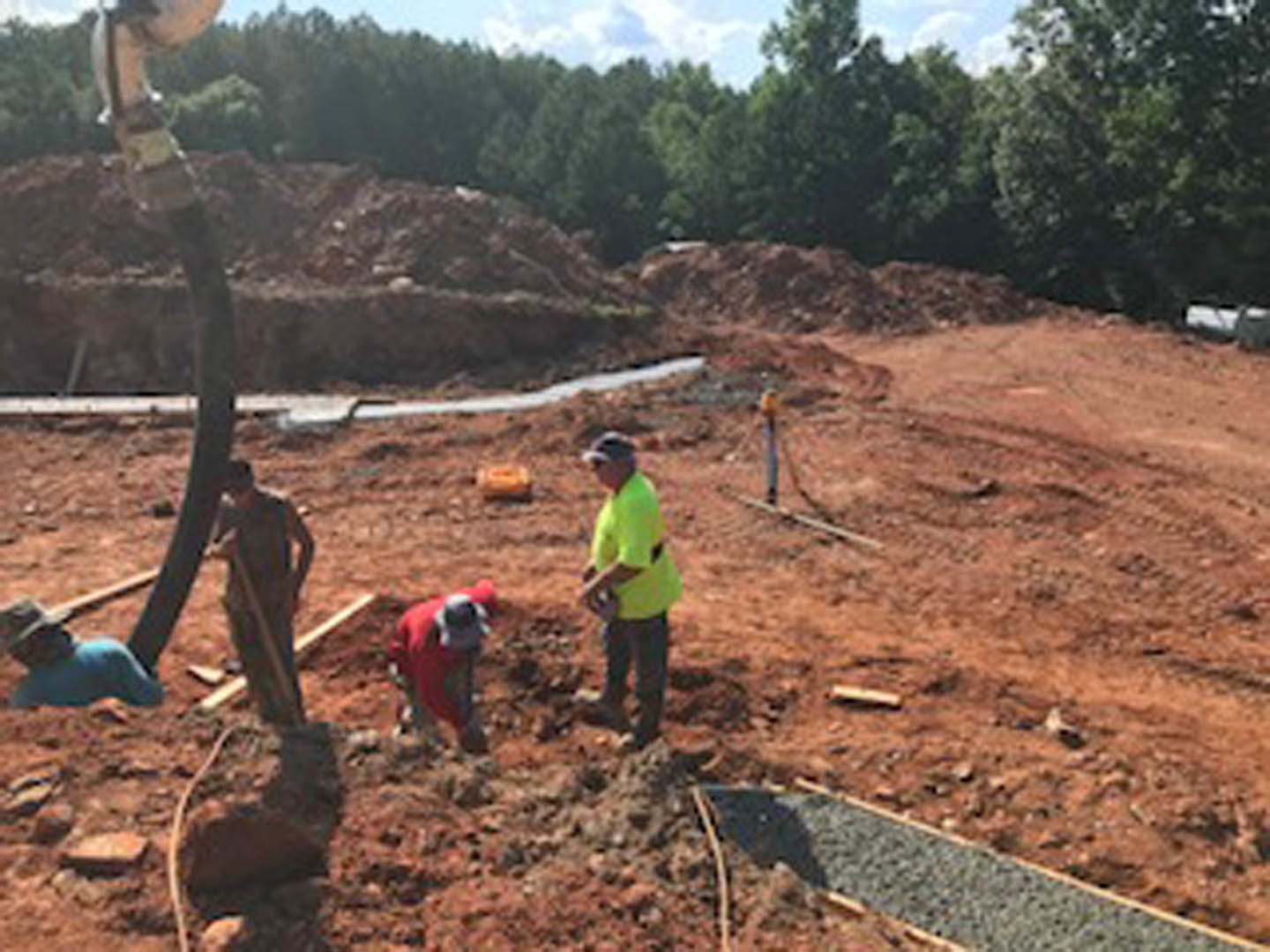 Framed custom home exterior under construction with workers in safety gear, muddy ground, scattered building materials, and trees in the background