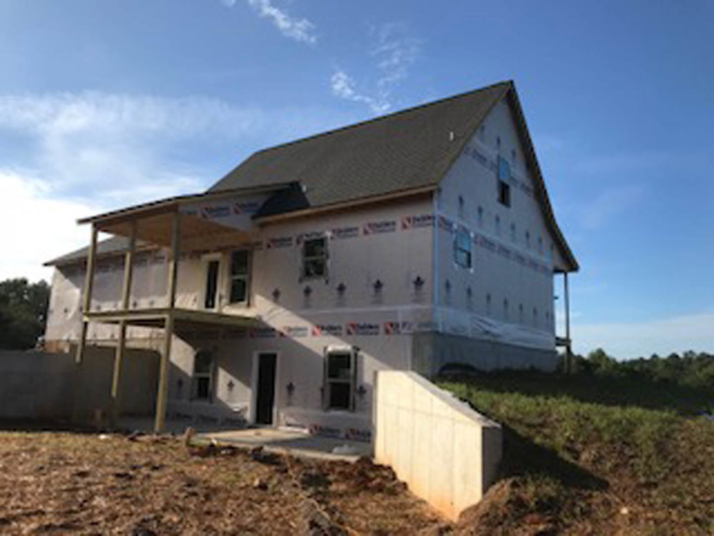 Concrete exterior wall and unfinished porch on custom home construction site, surrounded by dirt lot and trees under cloudy sky