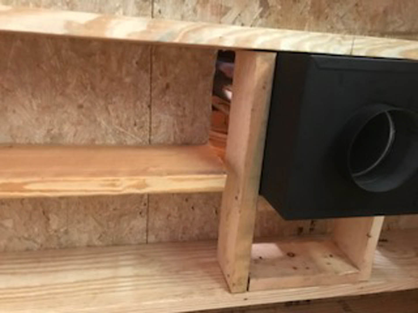Black loudspeaker with circular opening sits on light plywood shelf in modern interior.