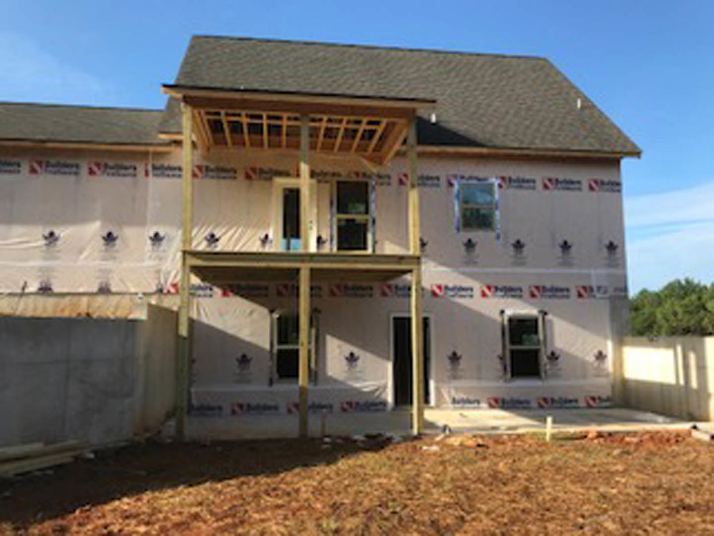 Wood-framed house under construction with covered porch, exposed beams, unfinished roof, and grassy yard in foreground