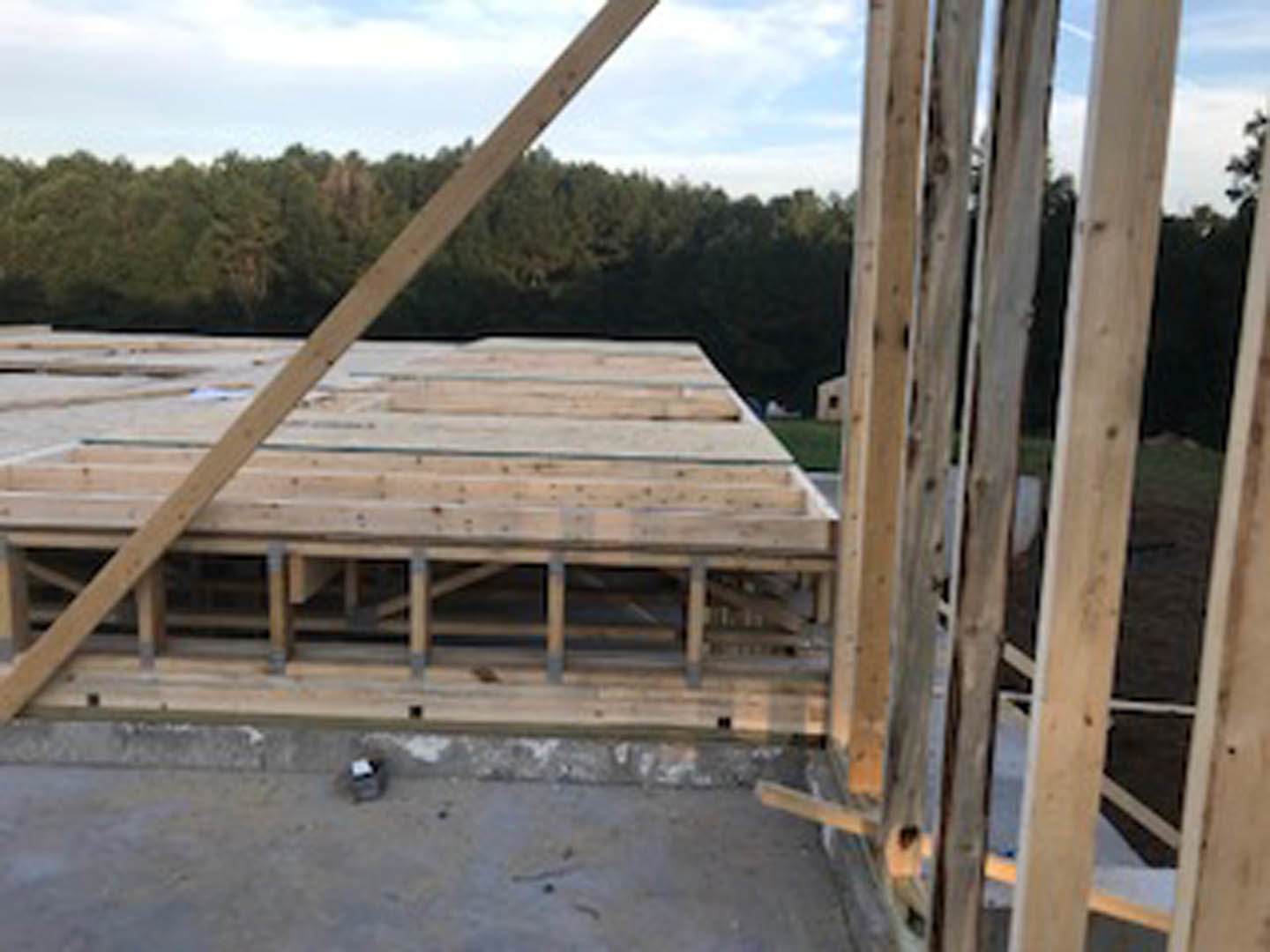 Wooden framing structure of a house under construction with exposed beams and planks, surrounded by trees and cloudy sky in the background