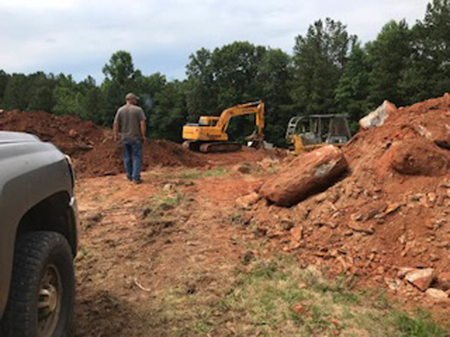 Bulldozer parked beside large mound of soil on construction site, man standing in front, trees and blue sky in background