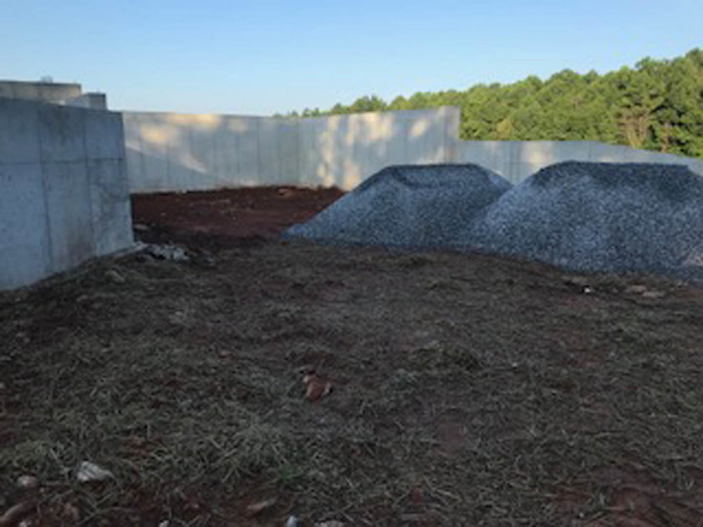 Gravel pile on bare dirt field bordered by white fence, blue sky with scattered clouds overhead, trees and grass in background